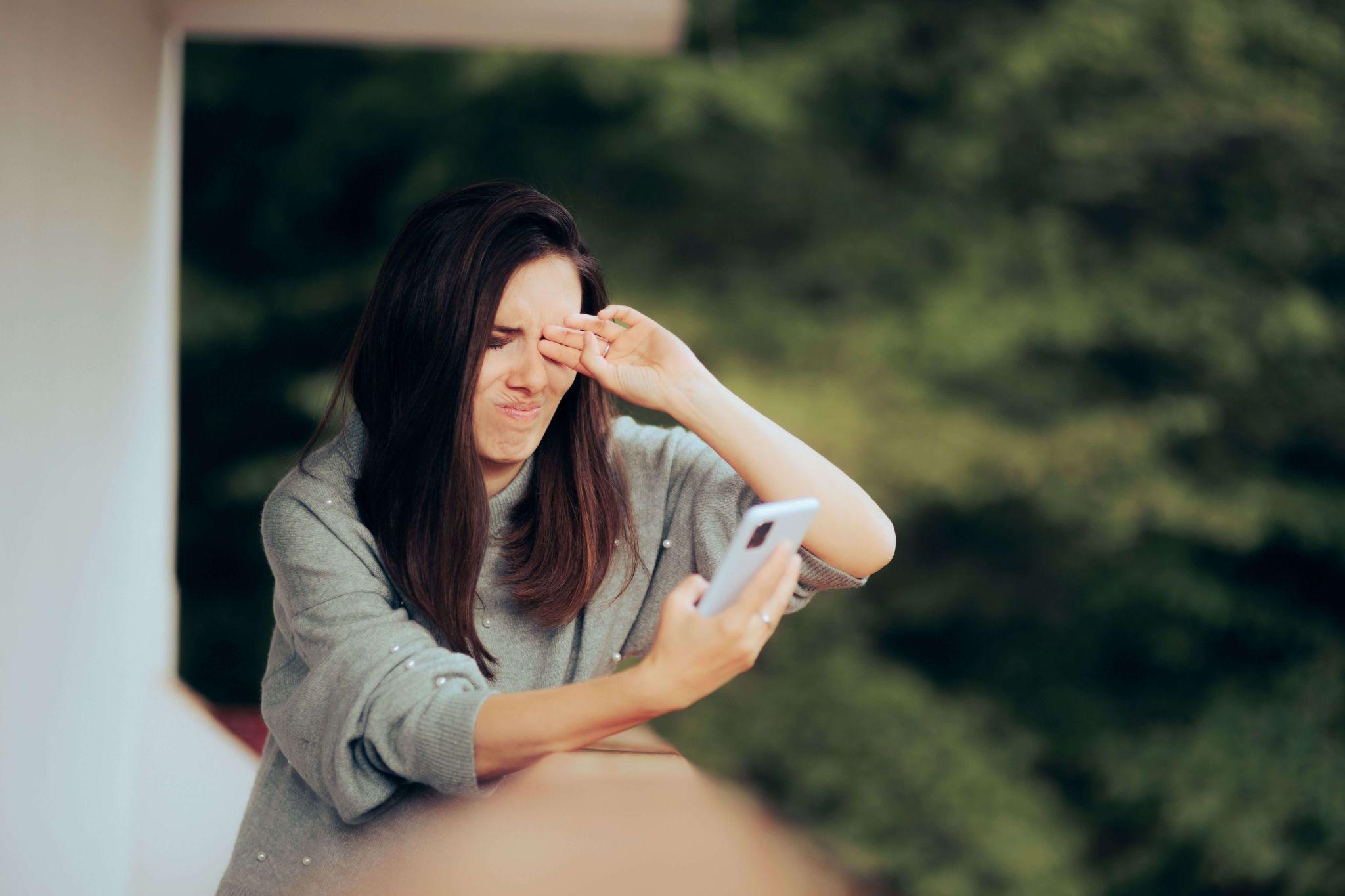 A person rubs their eyes and squints in discomfort from looking at their phone and being outside in bright light after an eye dilation exam.