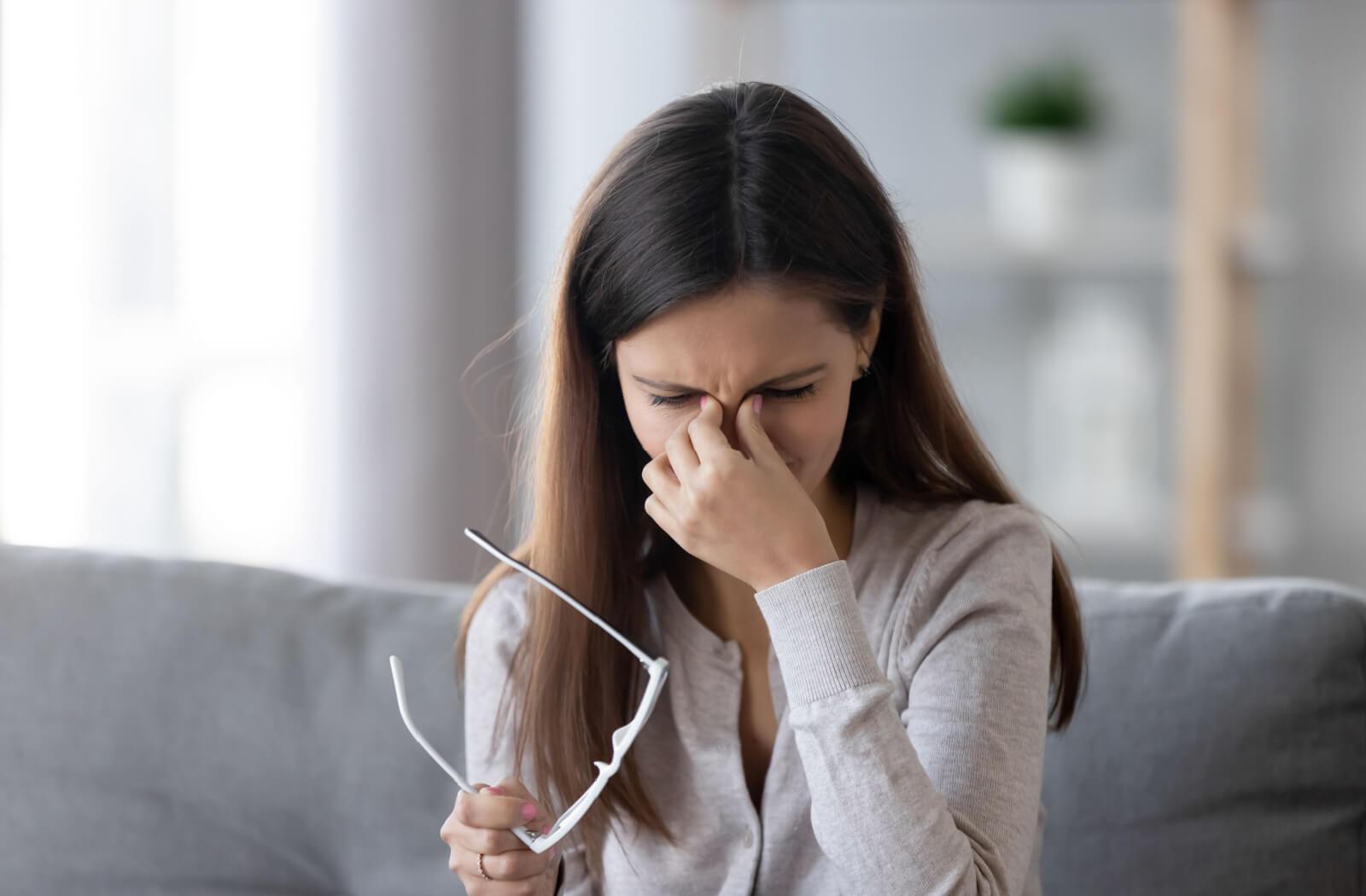 A young adult sitting on their couch at home, taking off their glasses and rubbing their eyes due to dry eye discomfort.