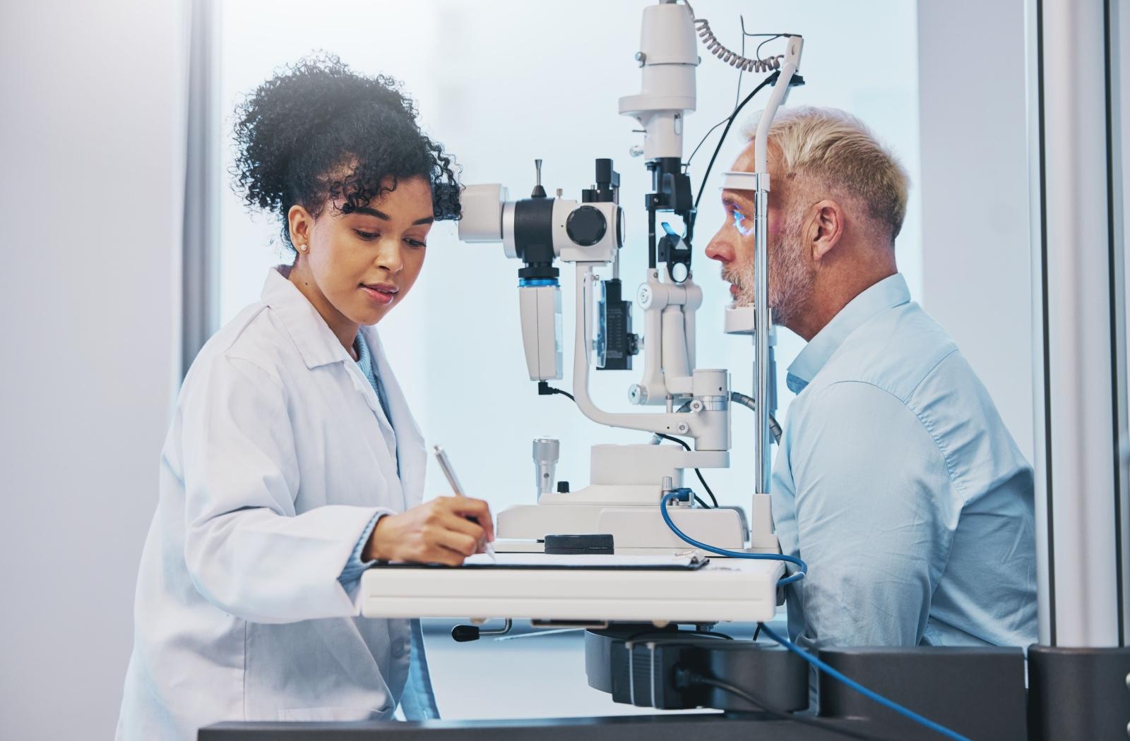 Patient undergoing an eye exam with their head positioned in a diagnostic machine, while the optometrist writes a note.